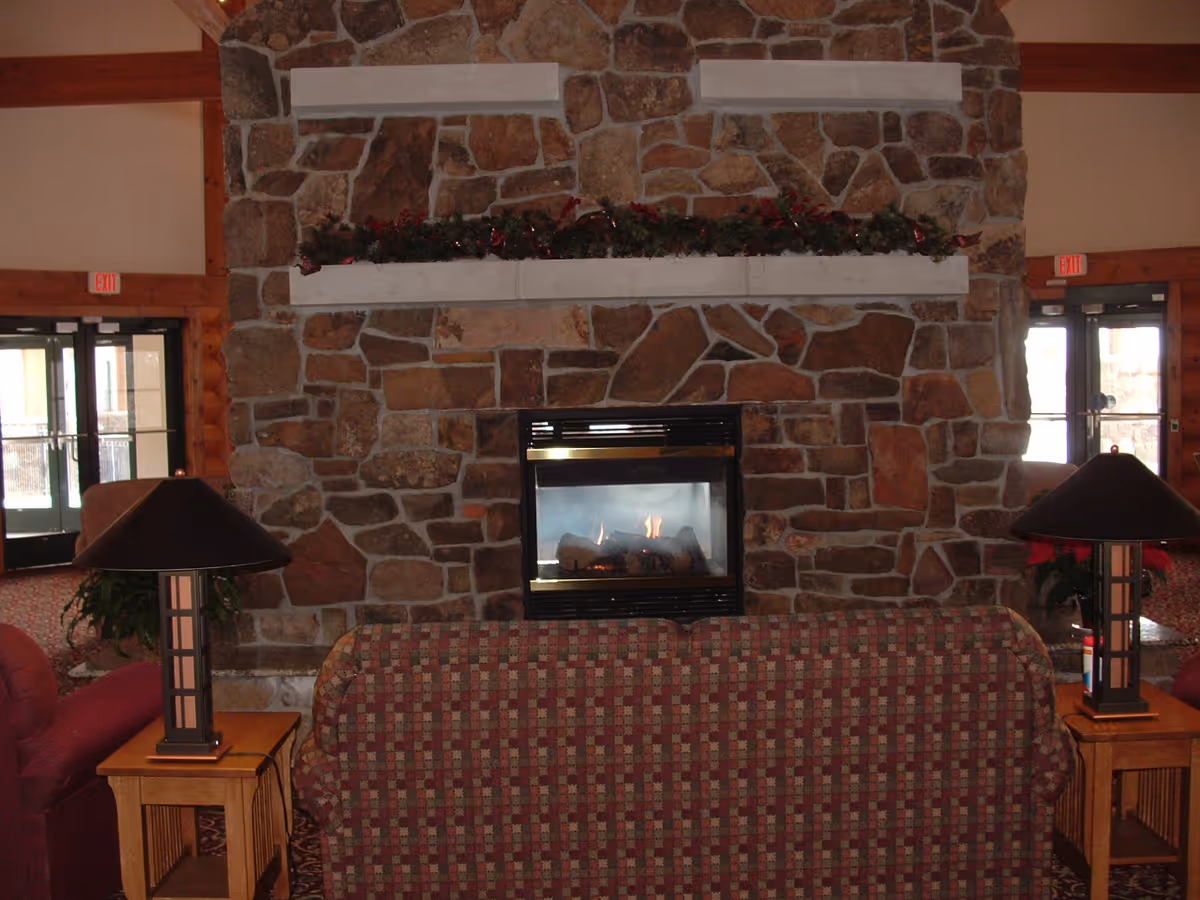 Cozy living room area with a stone fireplace in the center, decorated with garland on the mantel. In front of the fireplace is a patterned sofa flanked by two wooden side tables, each with a lamp. Double glass doors are visible on both sides of the fireplace.