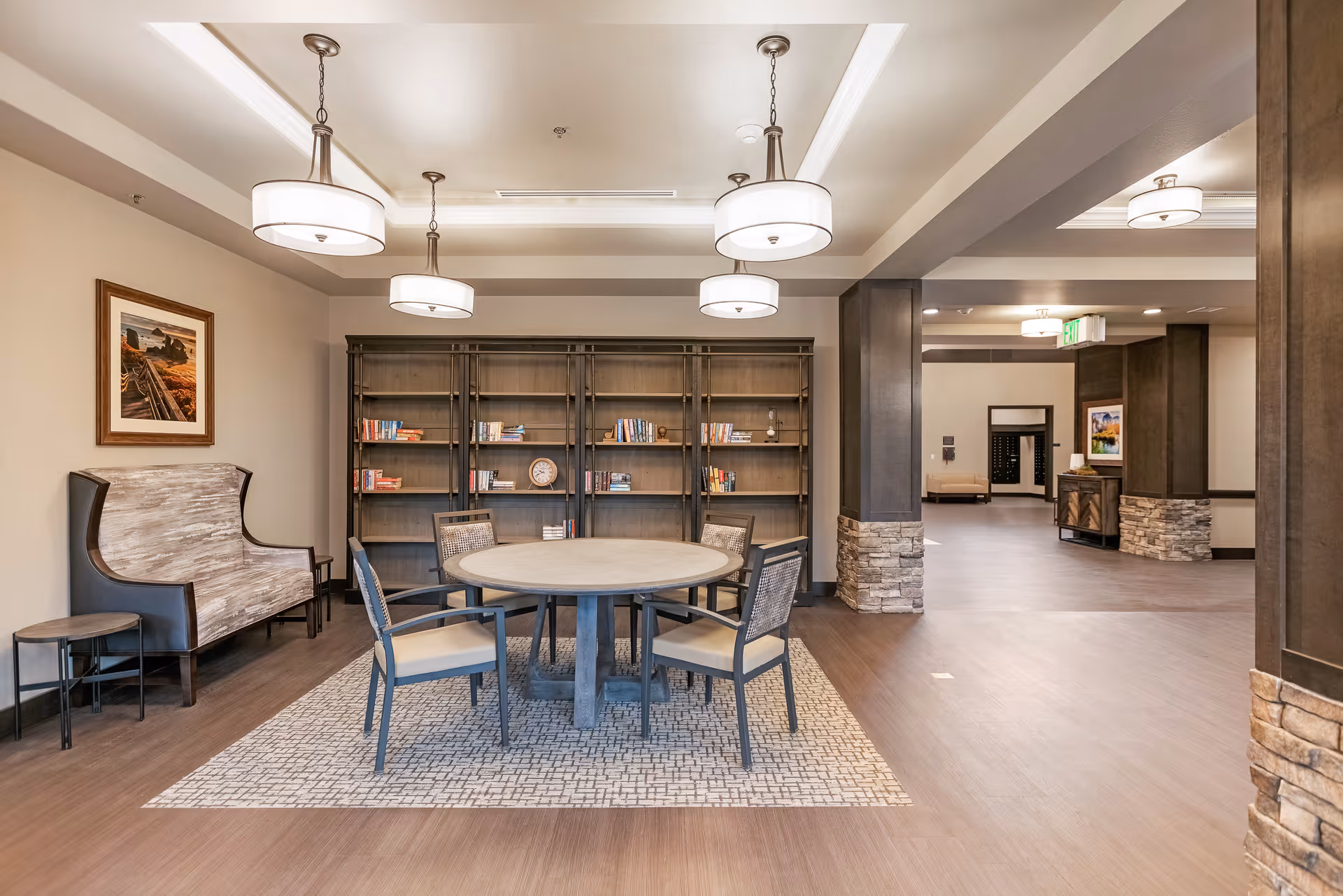 Bright communal lounge with a round table and chairs on a rug, bookshelves along the back wall, and seating under pendant lights.