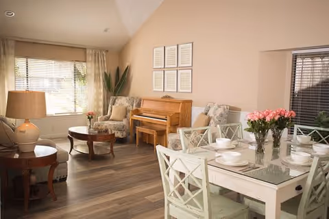 A cozy and well-lit living and dining area with a wooden piano against the wall, two cushioned armchairs, a round wooden coffee table, a side table with a lamp, and a dining table set with white dishes and a vase of pink flowers. Large windows with blinds and curtains allow natural light to fill the room.