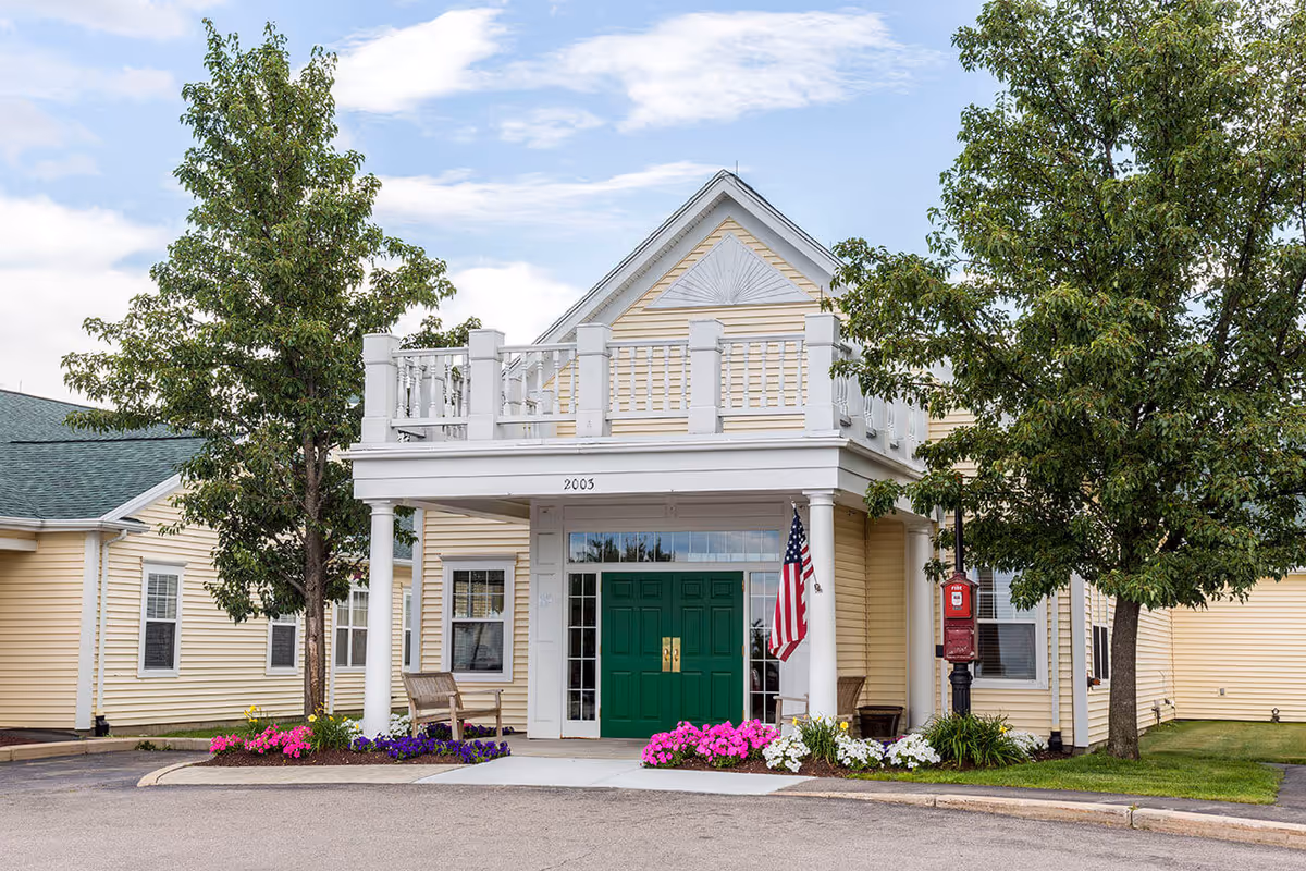 Front exterior view of a yellow building with white trim and a green double door entrance. There is a small porch with white columns and a balcony above it. Two trees flank the entrance, and colorful flowers are planted along the walkway. An American flag is displayed near the door.