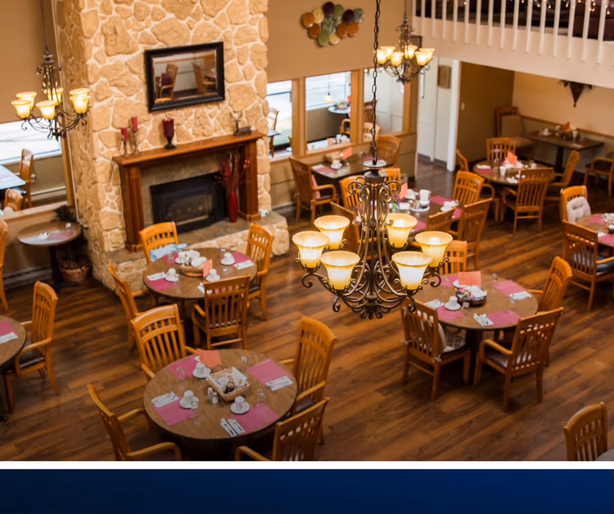 Interior view of a retirement facility dining room with multiple round wooden tables set with cups, plates, and pink placemats. The room features wooden chairs, a stone fireplace with a wooden mantle, a mirror above the fireplace, and two ornate chandeliers hanging from the ceiling. The floor is wooden, and there are windows letting in natural light.