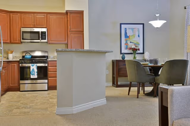 Interior view of a senior living facility showing a kitchen with wooden cabinets, a stainless steel stove and microwave, and a small island counter. Adjacent to the kitchen is a dining area with a round table, four green upholstered chairs, a sideboard with decorative items, and a framed abstract painting on the wall.