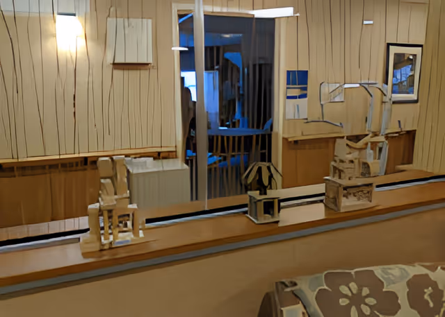 Interior view of a room with wooden panel walls, a large mirror reflecting part of the room, and a shelf holding three small decorative wooden sculptures. A patterned cushion is partially visible in the foreground.