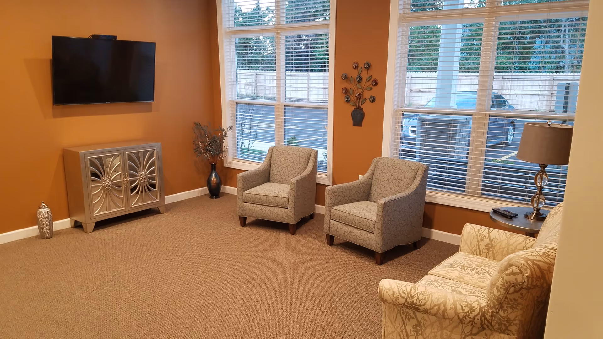 A cozy living room area with two patterned armchairs and a floral upholstered sofa arranged near large windows with white blinds. A flat-screen TV is mounted on an orange wall above a decorative silver cabinet. A small side table with a lamp and a vase with dried flowers are also visible.