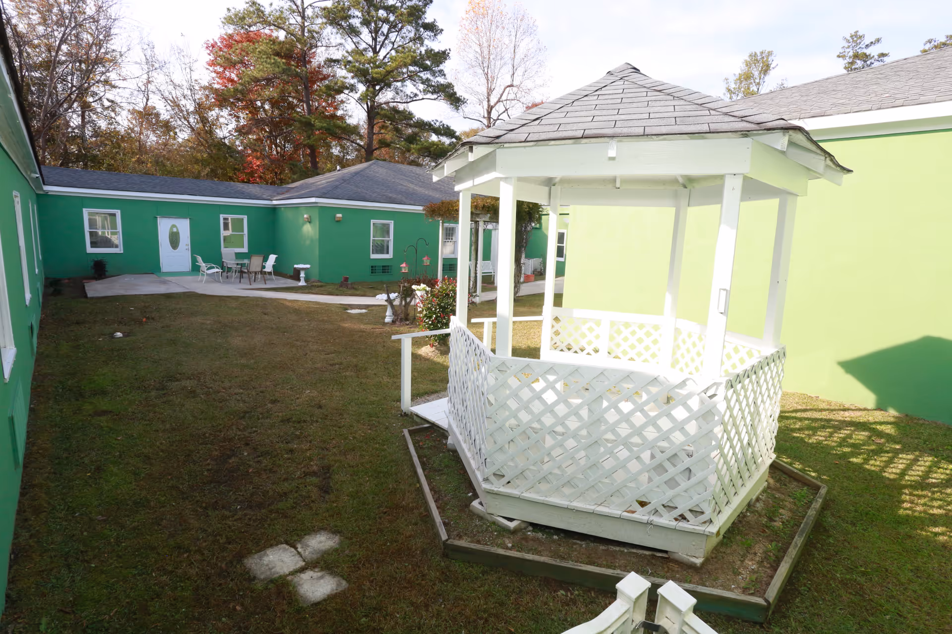 Outdoor courtyard area of St. Francis Assisted Living Community featuring a white wooden gazebo with lattice sides, surrounded by green buildings with multiple windows and a door. There is a small patio with a table and chairs, some bird feeders, and trees with autumn foliage in the background.