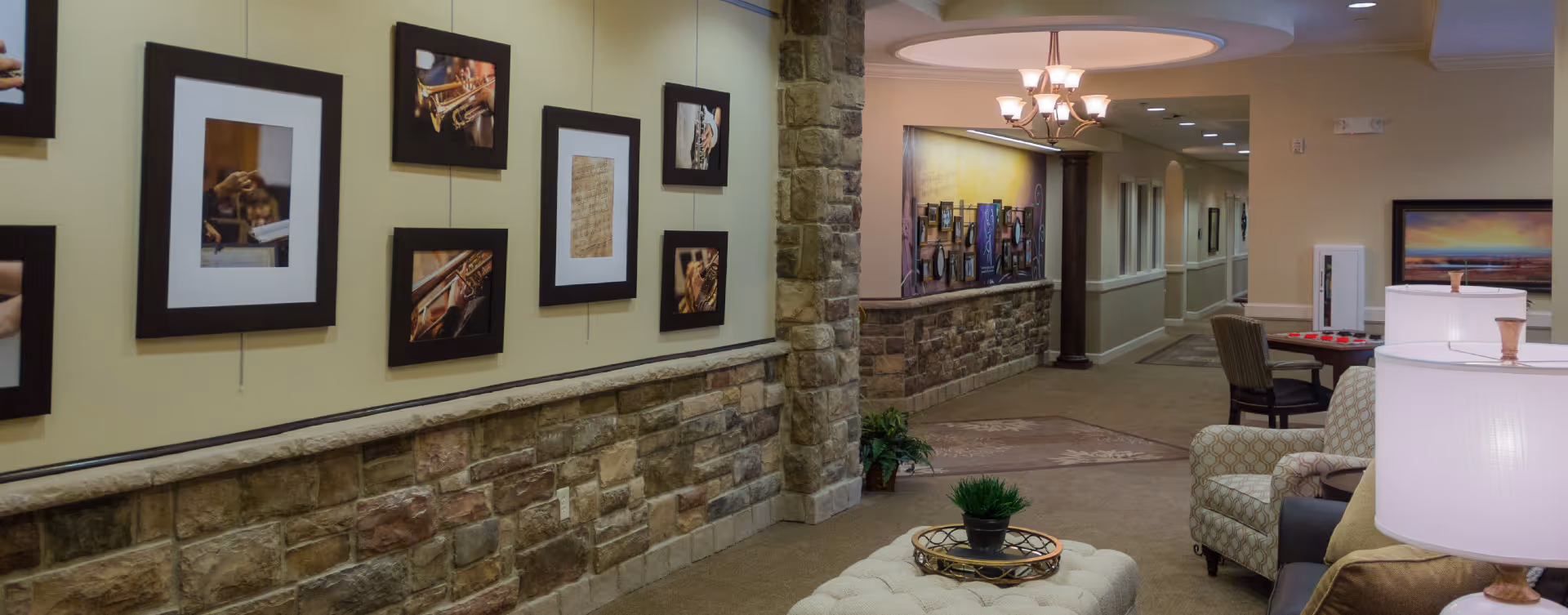 Interior hallway of a senior living facility with stone half-walls and beige walls. The left wall is decorated with framed pictures of musical instruments and sheet music. The hallway leads to a seating area with patterned armchairs, a tufted ottoman with a small plant on a tray, and table lamps. There is a chandelier on the ceiling and additional framed artwork on the walls.