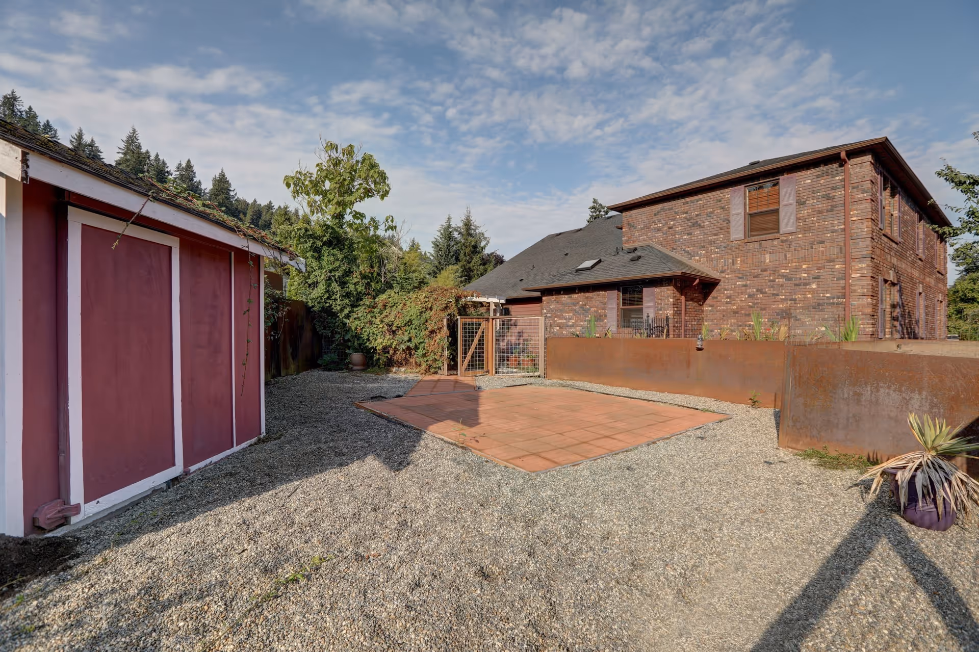 Outdoor view of a gravel yard area with a red shed on the left and a brick two-story building on the right. There is a small fenced gate and a tiled square patio area in the center. Trees and bushes are visible in the background under a partly cloudy sky.