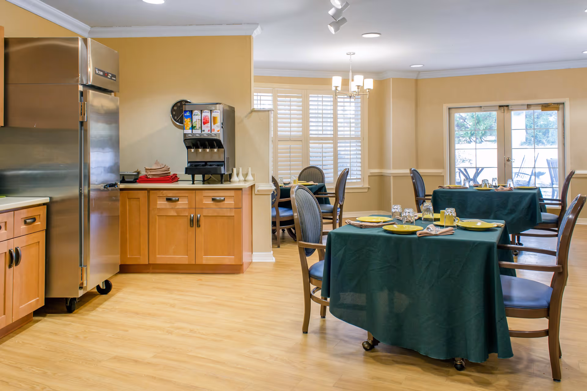 A dining room with wooden floors and beige walls featuring several tables covered with dark green tablecloths. Each table is set with yellow plates, glasses, and napkins. There are wooden chairs with blue cushions around the tables. In the background, there is a beverage dispenser on a wooden cabinet, a large stainless steel refrigerator, and windows with white shutters. Double glass doors lead to an outdoor patio area.