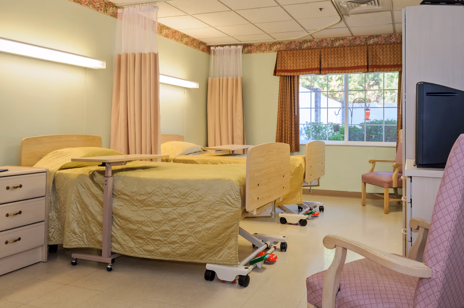 A room with two hospital-style beds covered with yellow quilts, each equipped with an overbed table. The beds have privacy curtains hanging from the ceiling. There is a window with brown patterned curtains letting in natural light. The room also contains a wooden dresser, a TV on a stand, and two upholstered chairs with wooden arms.