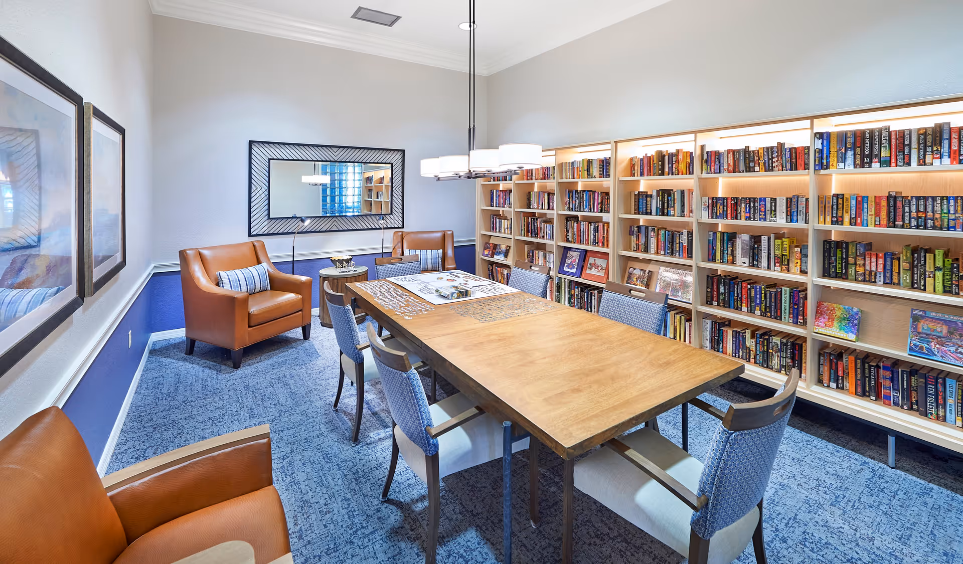 A bright and cozy reading and activity room with a large wooden table in the center surrounded by six chairs. The table has a partially completed jigsaw puzzle on it. Along one wall, there are tall bookshelves filled with books and some framed pictures. Two comfortable brown leather armchairs with striped pillows are placed near the wall with a large rectangular mirror above them. The room has blue carpet flooring and light-colored walls with blue wainscoting.