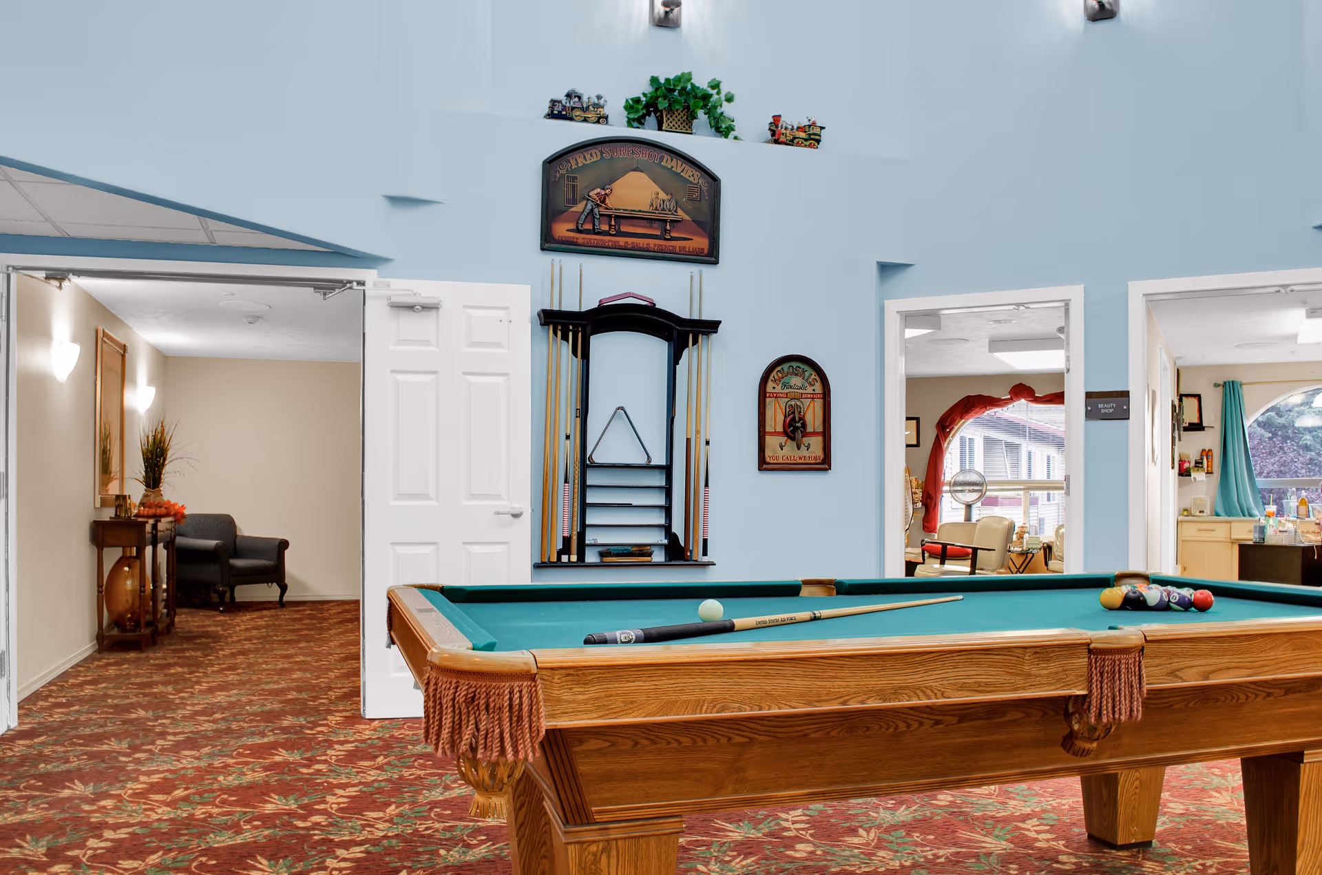 Interior view of a recreational room with a pool table in the foreground. The pool table has a cue stick and balls arranged on it. Behind the pool table, there is a wall-mounted rack holding pool cues and a triangle rack. The room has light blue walls and a patterned carpet with red and green tones. Two open doorways lead to adjacent rooms, one with a chair and table, and the other with a window and additional seating. Decorative signs and small plants are placed above the pool cue rack.