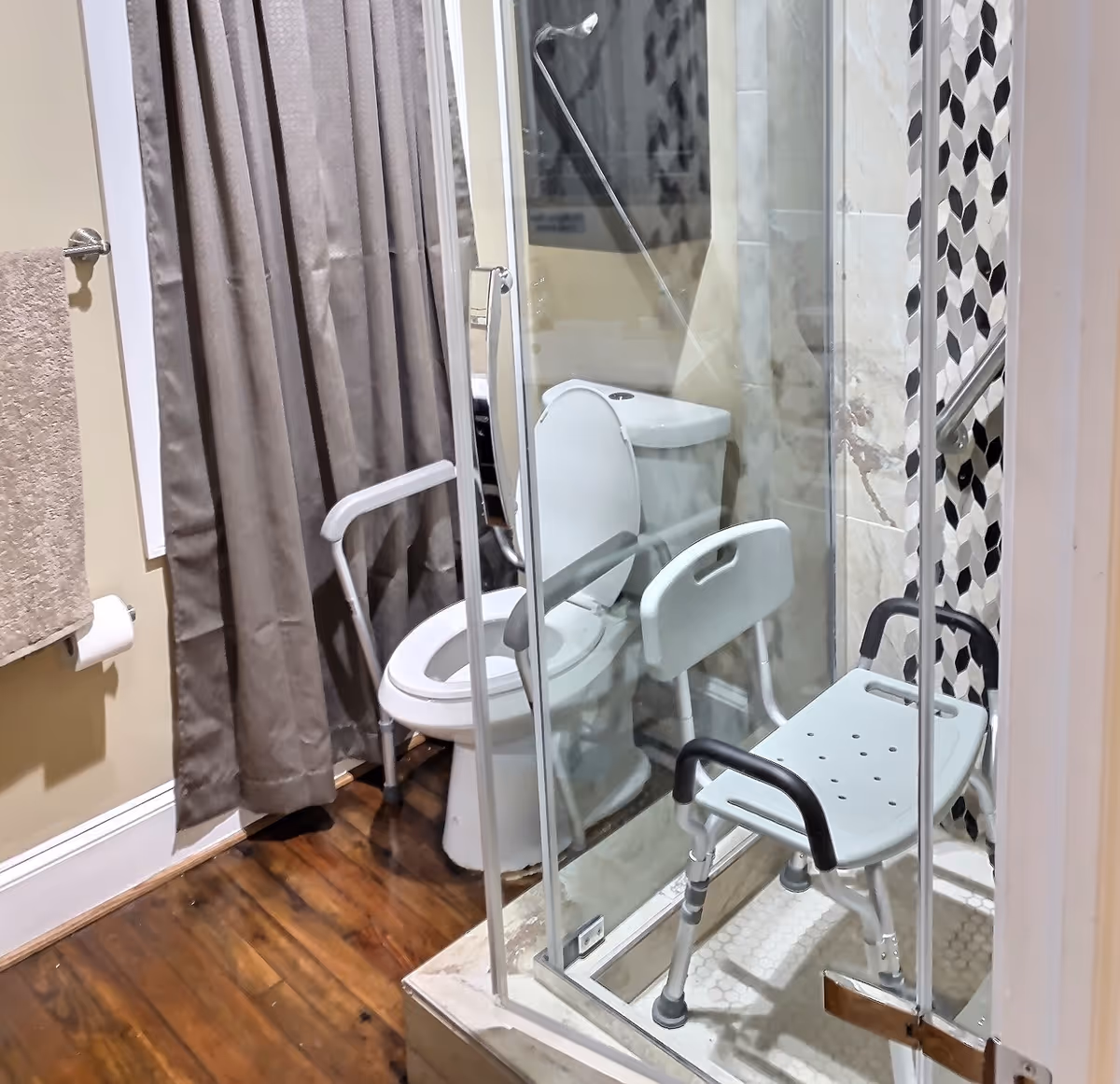 Bathroom showing a toilet with safety rails and a glass-enclosed shower containing a shower chair and grab bars.