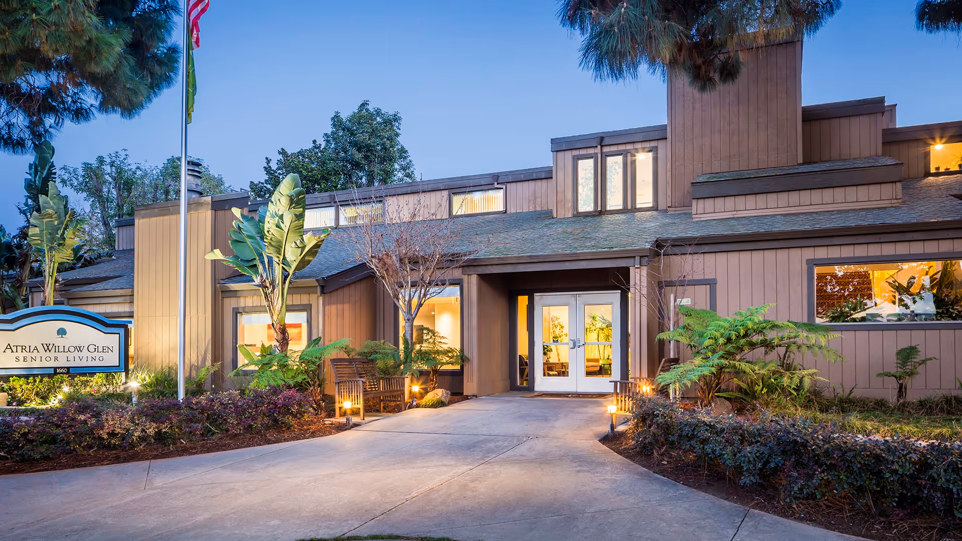 Exterior view of Atria Willow Glen senior living facility at dusk, showing the entrance with double doors, surrounding landscaping with plants and trees, a bench, and a sign with the facility name.