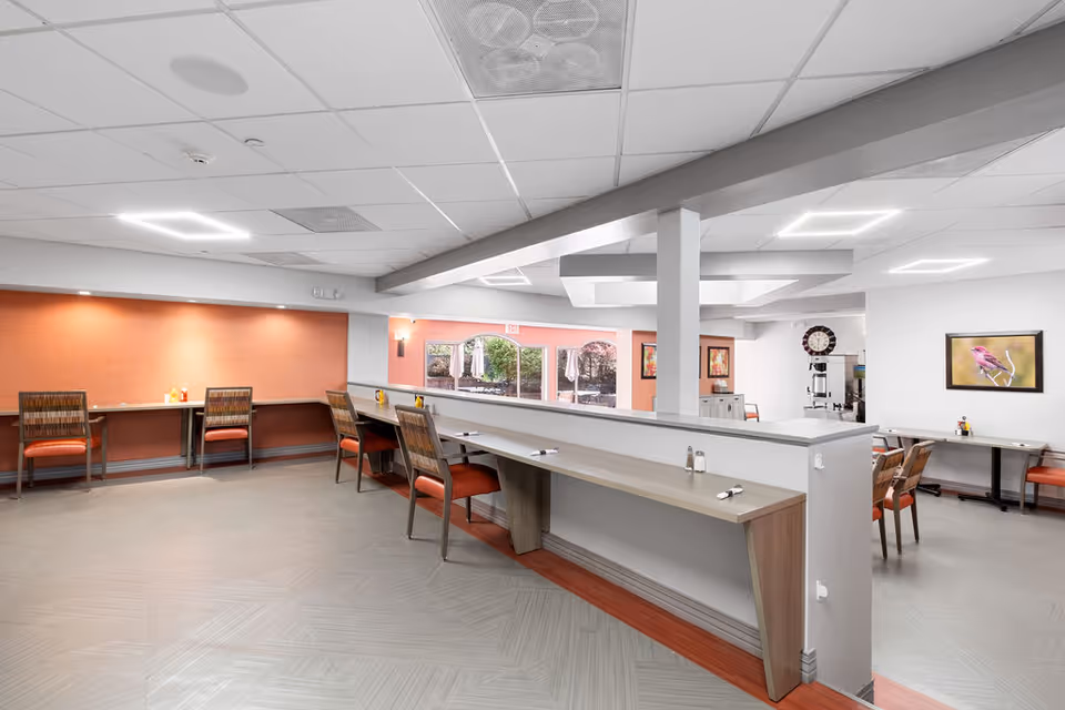 Open, well-lit communal dining area with counter seating, tables and chairs adjacent to large windows.