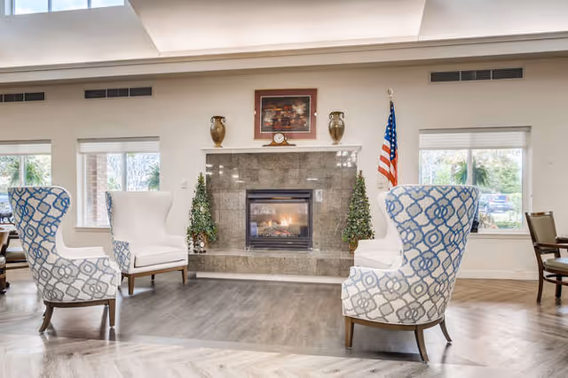 A cozy senior living community lounge area featuring a fireplace with a marble surround, two decorative vases, a clock, and a framed picture above it. Two patterned armchairs and two white armchairs are arranged around the fireplace on a wood floor. Large windows on either side let in natural light, and an American flag stands to the right of the fireplace.
