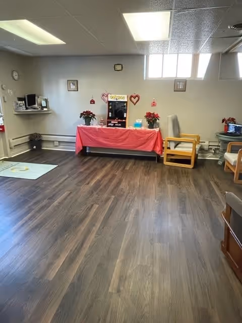 A well-lit room with wood flooring and white walls featuring a table covered with a red cloth, decorated with heart-shaped ornaments and potted plants. There are chairs along the walls, a small TV on a stand, a computer on a wall-mounted shelf, and a clock above the table. The room has a window near the ceiling letting in natural light.