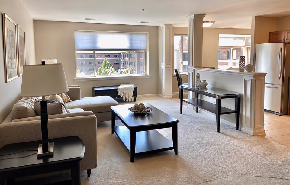 Sunlit open-plan living room featuring a beige sectional sofa, dark wood coffee table, and a partial view of the kitchen and large window.