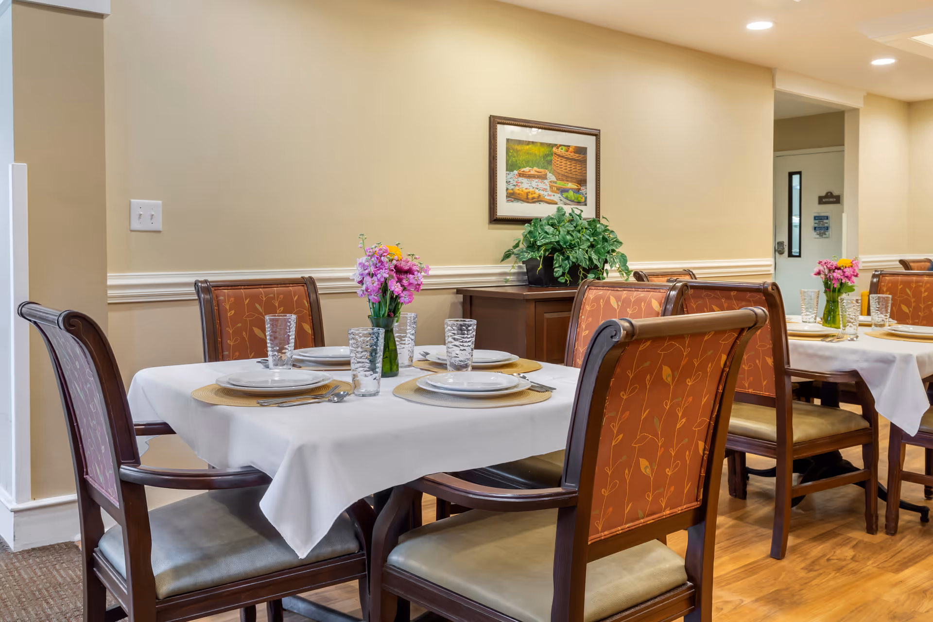 A dining area in Brookdale Chapel Hill with tables set for four, each covered with white tablecloths, plates, glasses, and silverware. The chairs have wooden frames with orange patterned upholstery. There are small flower arrangements in green vases on the tables, and a framed picture and a potted plant are visible on a wooden sideboard against a beige wall.