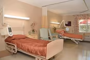 A senior care patient room with two adjustable hospital beds, a nightstand, chairs, and a window with curtains.