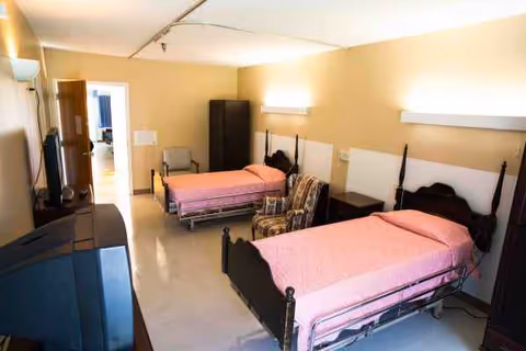 Two pink beds with nightstands and a small seating area in a nursing home bedroom.