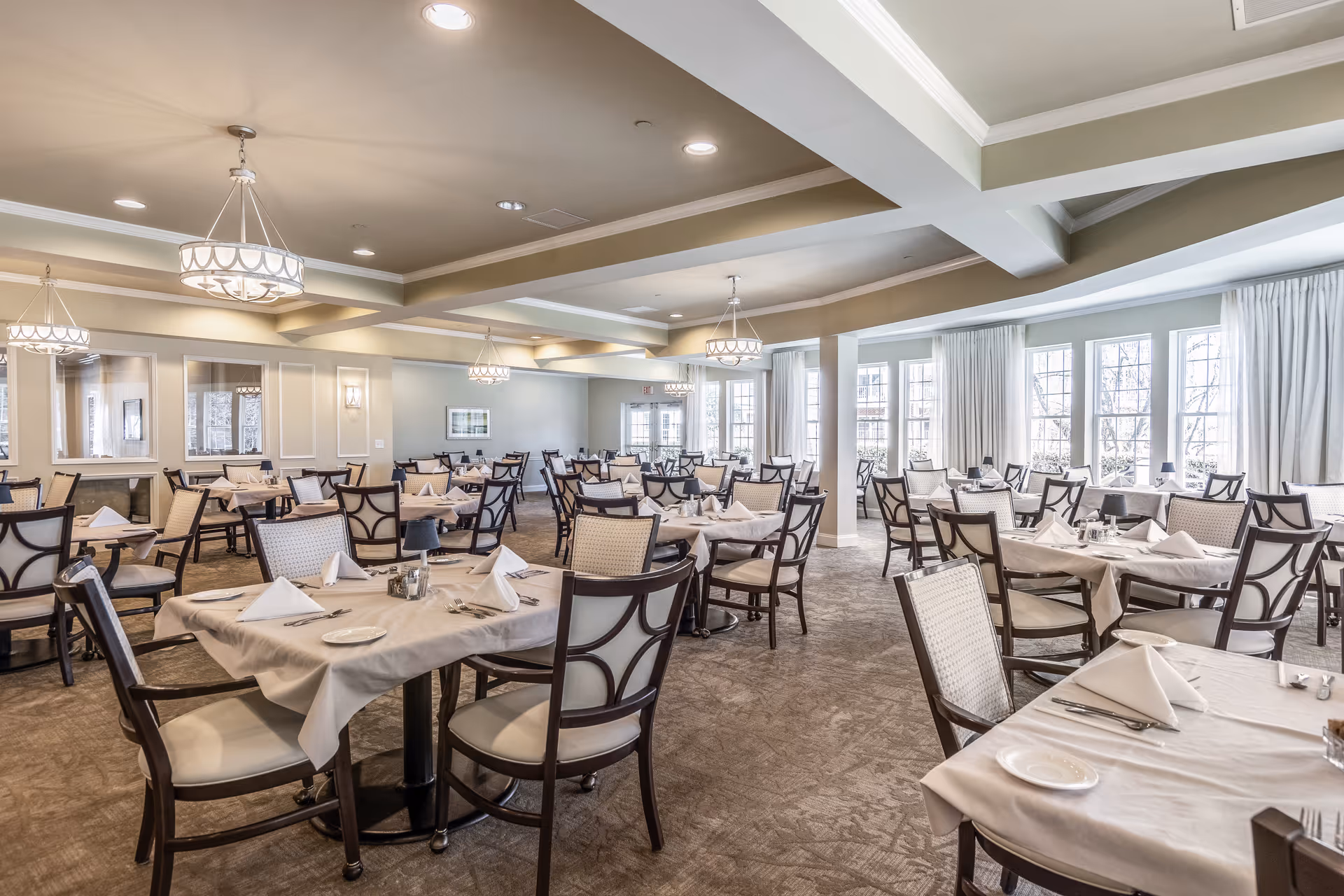 A spacious and well-lit dining room with multiple tables covered in white tablecloths, each set with folded napkins, plates, and silverware. The room features large windows with white curtains, beige walls, and elegant ceiling light fixtures.