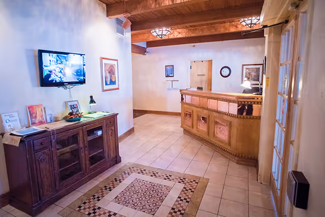 Interior view of a reception area with a wooden front desk, tiled floor with a decorative rug, a wooden cabinet with a lamp and framed pictures, a wall-mounted TV, and framed artwork on the walls. The ceiling has exposed wooden beams and decorative light fixtures.