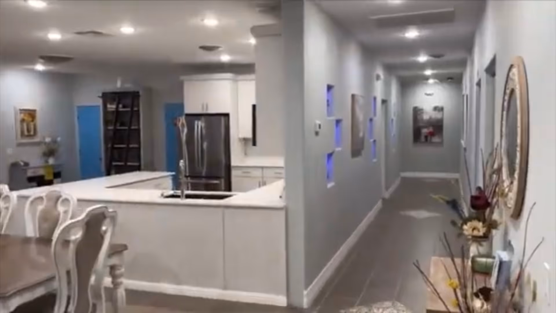 Open kitchen with a large island and sink next to a dining table, leading into a long decorated hallway.