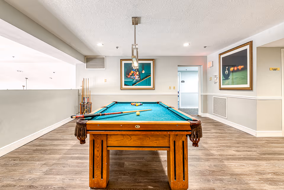 Interior view of a recreational room featuring a wooden pool table with a green felt surface. Pool cues rest on the table and a rack of cues is positioned against the wall. Two framed pictures of billiard balls hang on the walls. The room has wood flooring, white walls with a chair rail, and recessed ceiling lights.