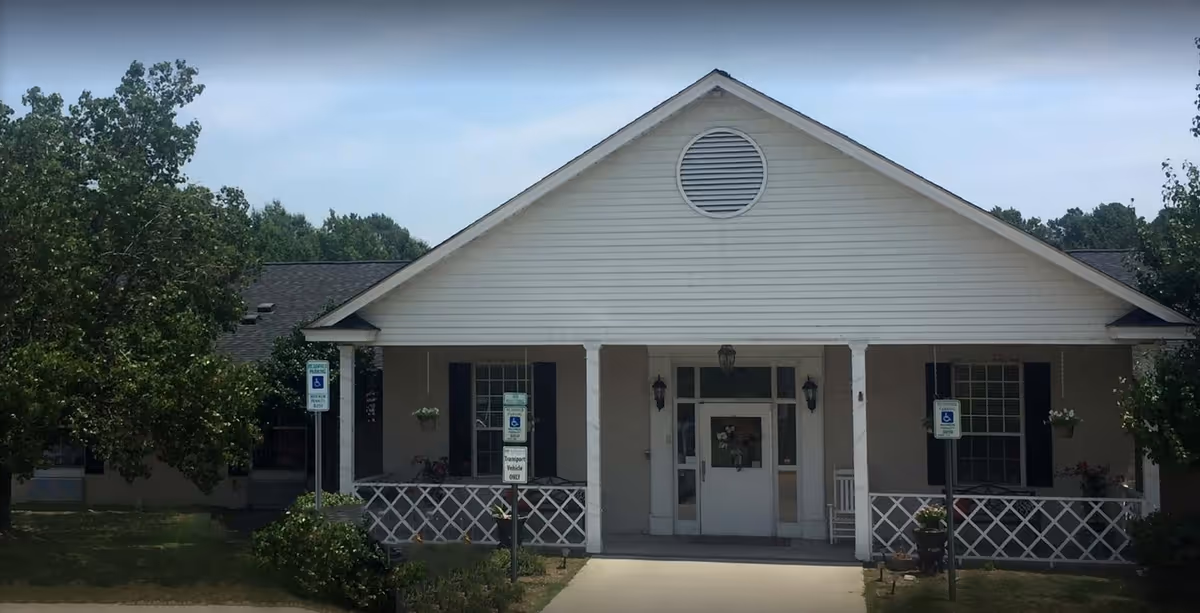 Front exterior view of a single-story building with a white facade, a peaked roof, and a covered porch supported by white columns. There are two handicap parking signs in front of the building, and greenery including trees and bushes surrounds the area.