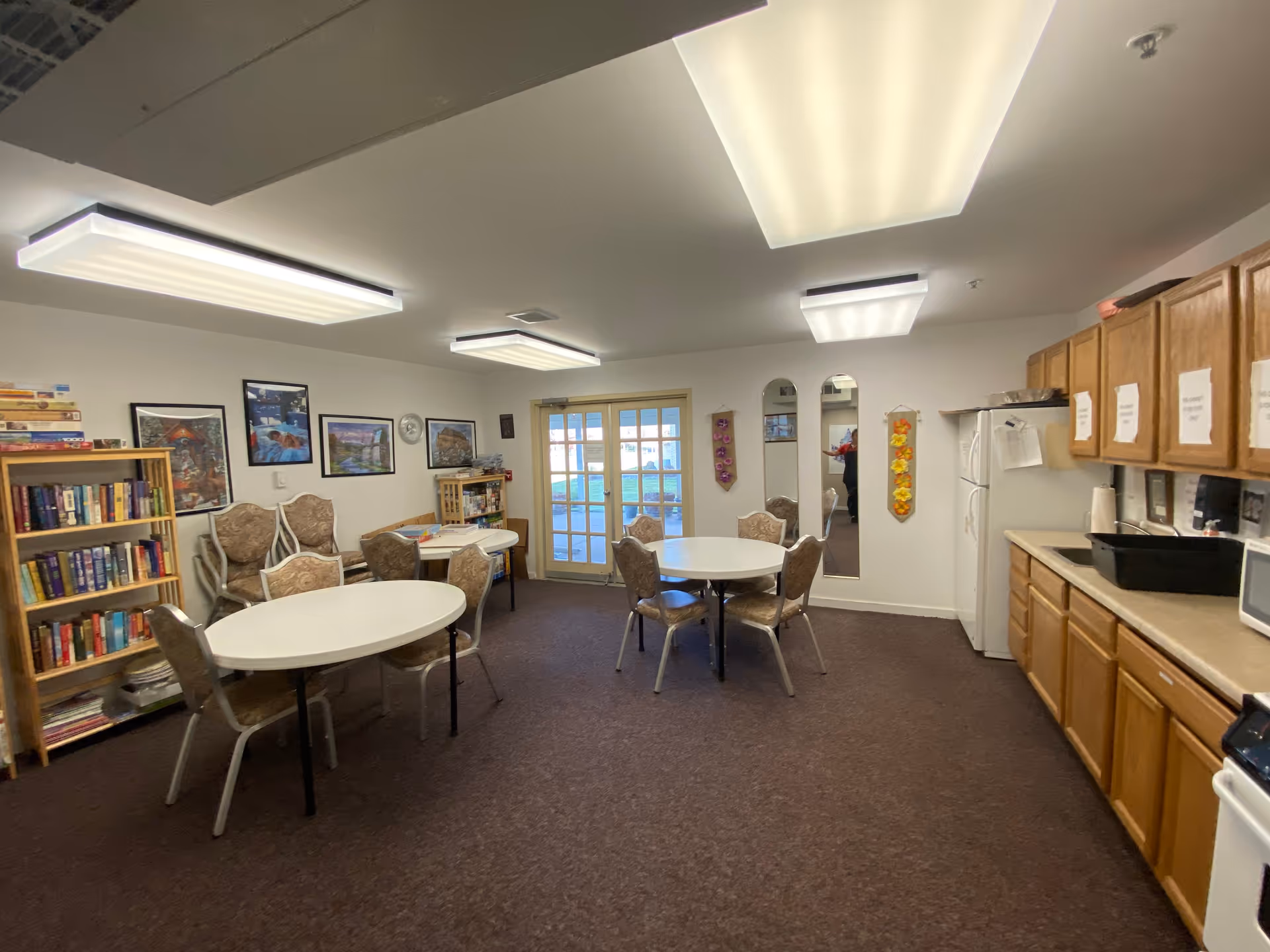A well-lit common room with round tables and chairs, a bookshelf filled with books and board games, and a kitchenette with wooden cabinets, a refrigerator, microwave, and sink. The room has carpeted flooring, white walls adorned with framed pictures and decorative wall hangings, and a glass door leading outside.