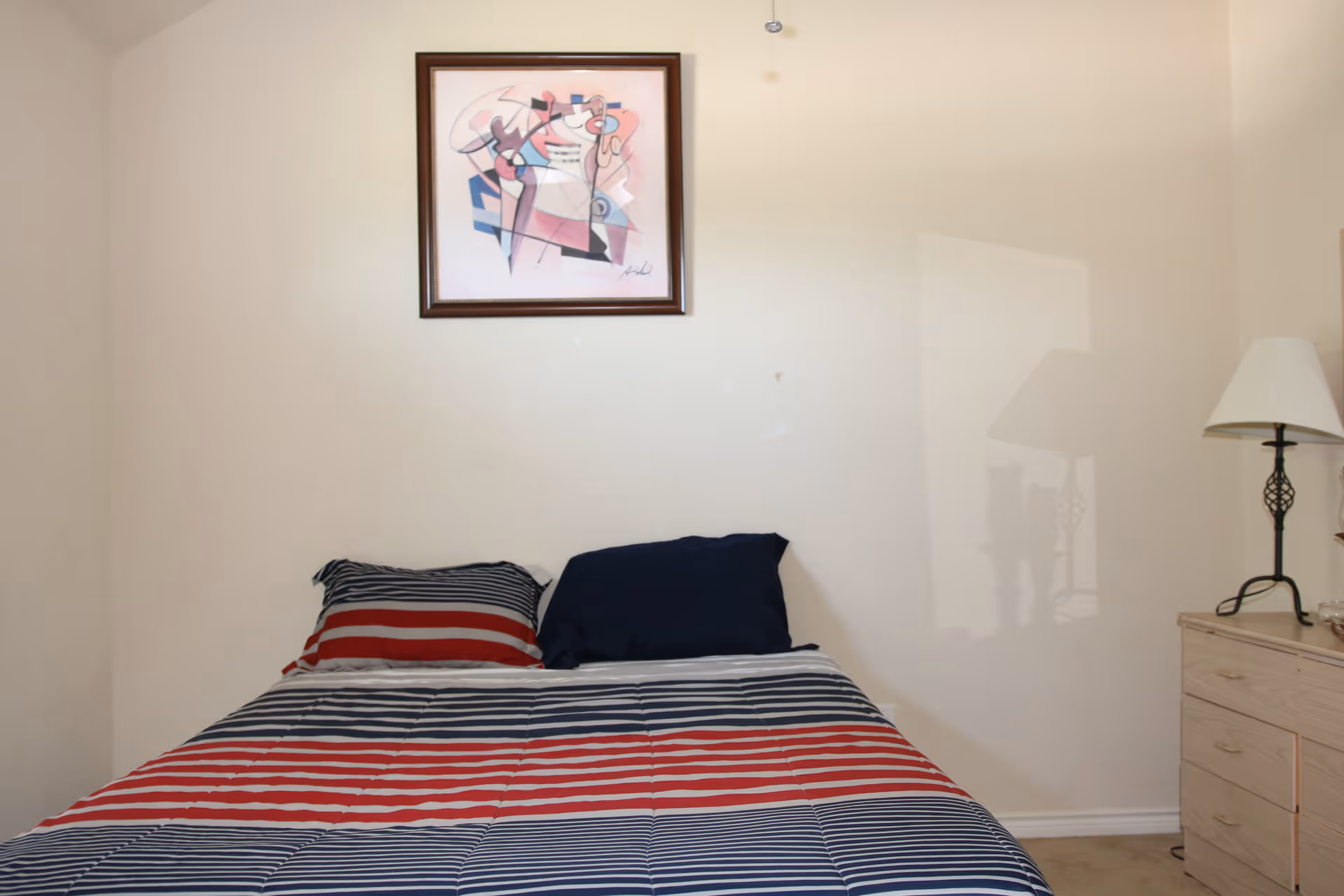 A bedroom with a bed covered in a striped comforter in red, white, and blue colors. There are two pillows on the bed, one with matching stripes and the other solid dark blue. Above the bed hangs a framed abstract painting. To the right of the bed is a wooden dresser with a decorative lamp on top. The walls are plain and light-colored.