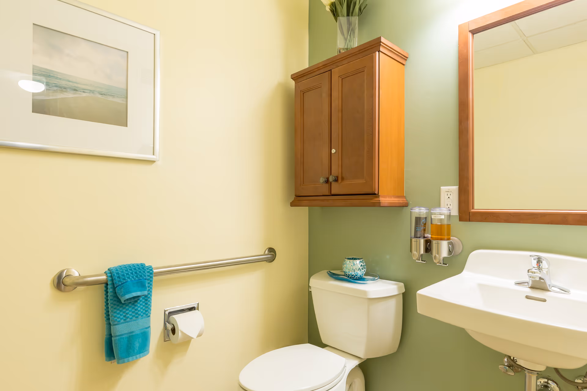 A clean and well-lit bathroom with a white toilet and sink. Above the toilet is a wooden cabinet with two doors and a vase with flowers on top. A large mirror with a wooden frame is mounted above the sink. On the wall next to the toilet, there is a stainless steel grab bar with a turquoise towel hanging on it and a toilet paper holder. A framed picture of a beach scene is hung on the wall.