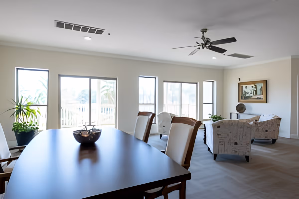 A bright and spacious room with large windows letting in natural light. In the foreground, there is a dark wooden dining table with a decorative bowl on it and cushioned chairs around it. In the background, there is a seating area with patterned armchairs, a side table, a plant, and a framed painting on the wall. A ceiling fan is mounted on the white ceiling.