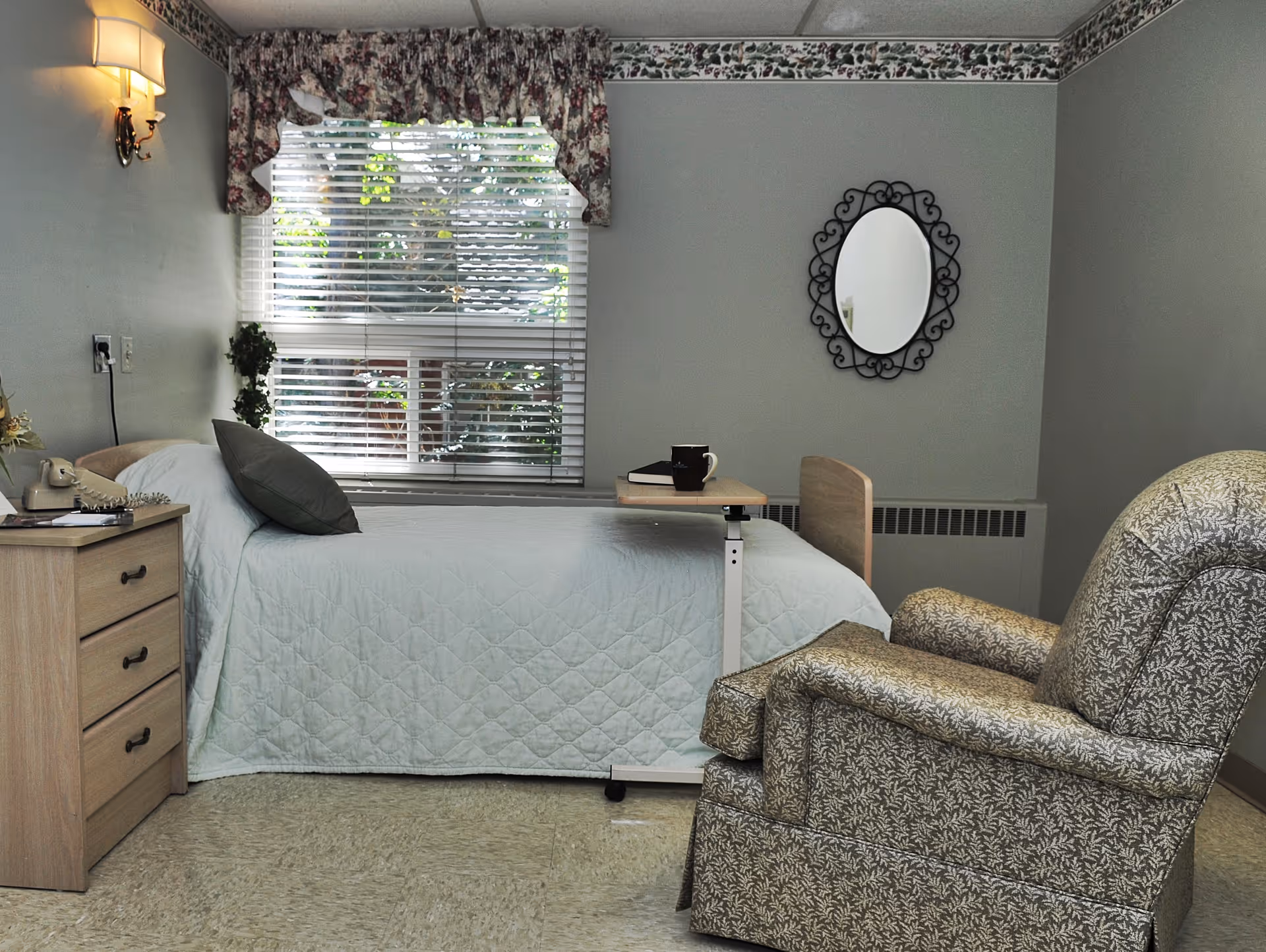 A tidy senior living bedroom with a single bed, bedside table, window with blinds, and an upholstered recliner.