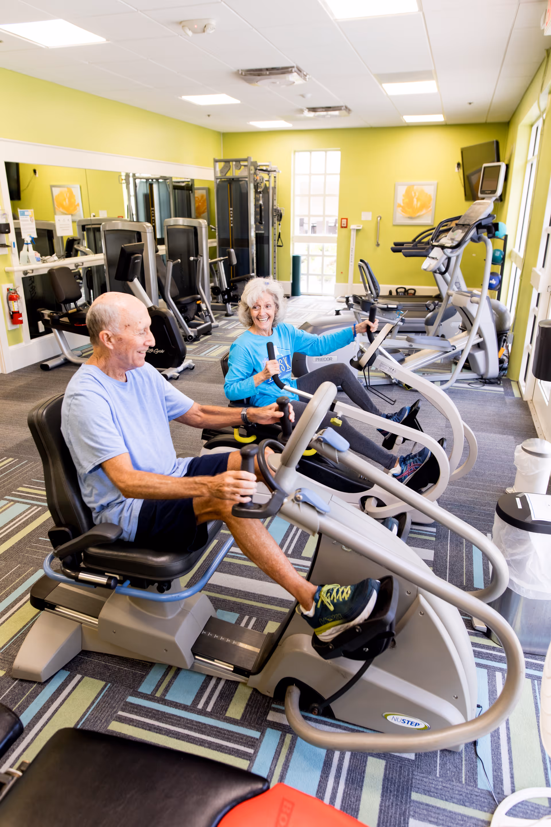 Two elderly individuals exercising on recumbent stationary bikes in a bright fitness room with green walls, various exercise equipment, and large windows letting in natural light.
