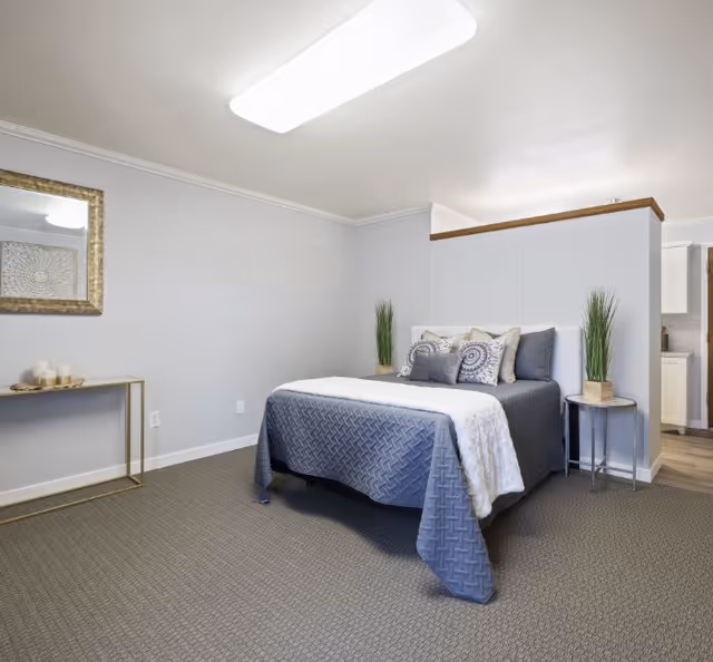 A neatly staged bedroom with a gray quilted bed, decorative pillows, side tables with potted plants, a console table and mirror against neutral walls.