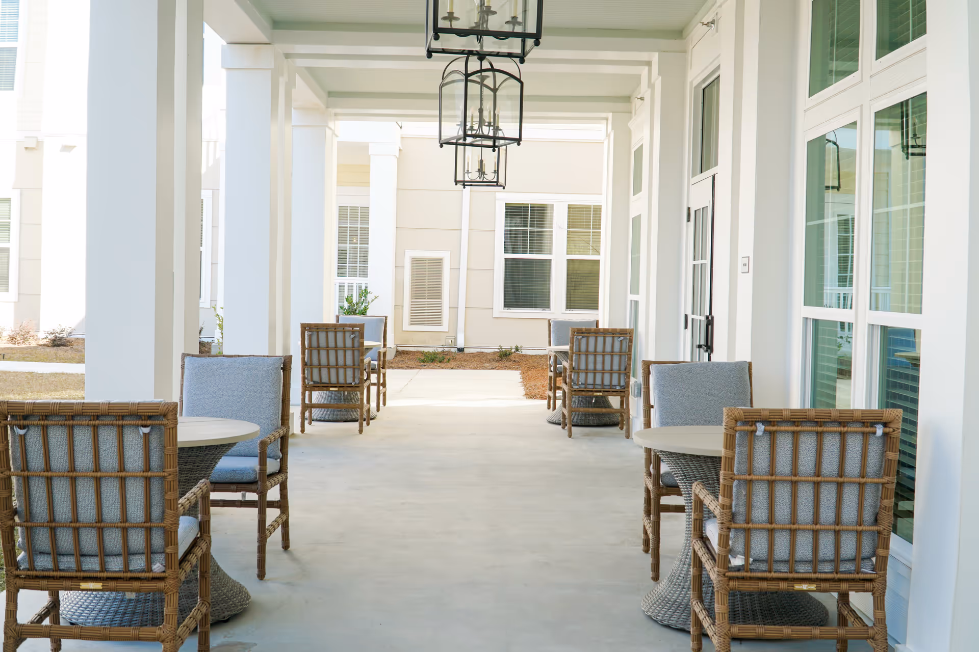 Covered outdoor patio area with multiple round tables and cushioned wicker chairs arranged in pairs along a walkway. The space is bright with white pillars and large windows on one side, and hanging lantern-style light fixtures overhead.