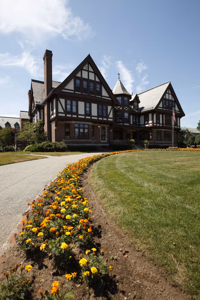 A large, traditional-style building with Tudor architectural elements, surrounded by a well-maintained lawn and a curved flower bed filled with yellow and orange flowers under a clear blue sky.