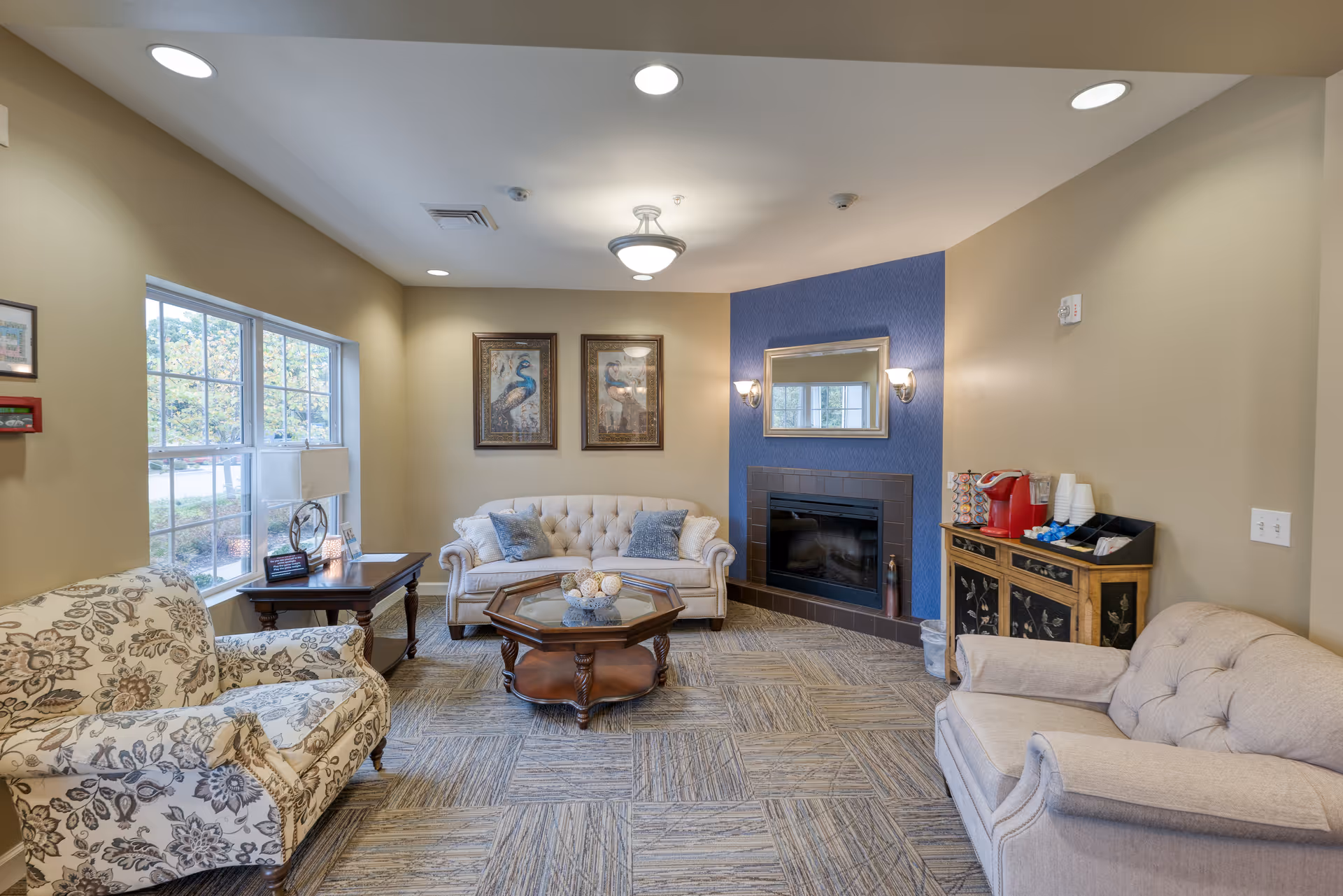 A cozy living room with beige walls and a blue accent wall featuring a fireplace. The room has a beige tufted sofa, a floral patterned armchair, and a beige armchair. A wooden coffee table with decorative items is centered on a patterned carpet. Two framed peacock pictures hang above the sofa. A window with a side table and lamp is on the left, and a small cabinet with a coffee maker and cups is on the right.