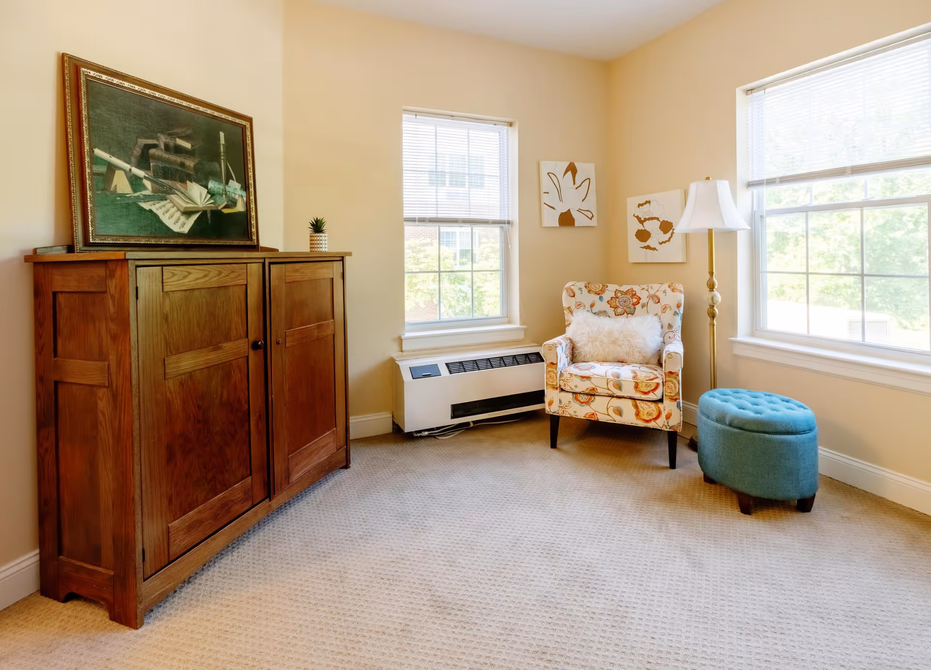 Bright sitting area with a floral armchair, blue ottoman, wooden cabinet, floor lamp, and two large windows.