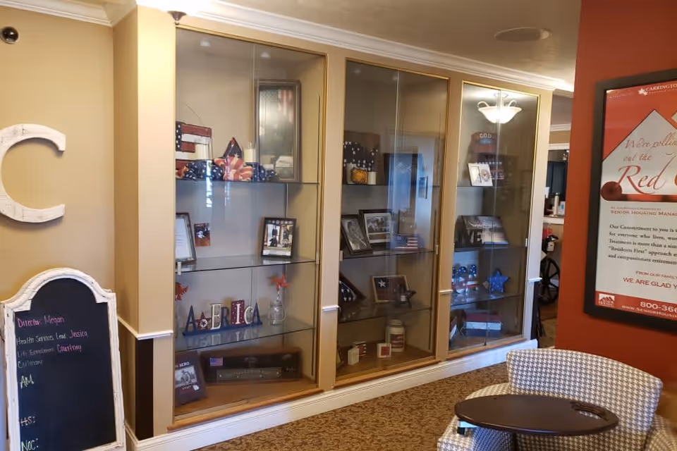 Interior view of a senior living facility showing a display cabinet with patriotic-themed decorations and memorabilia. To the left, there is a chalkboard with staff names written on it, and to the right, a red wall with a framed informational poster. In the foreground, there is a patterned chair with a small attached table.
