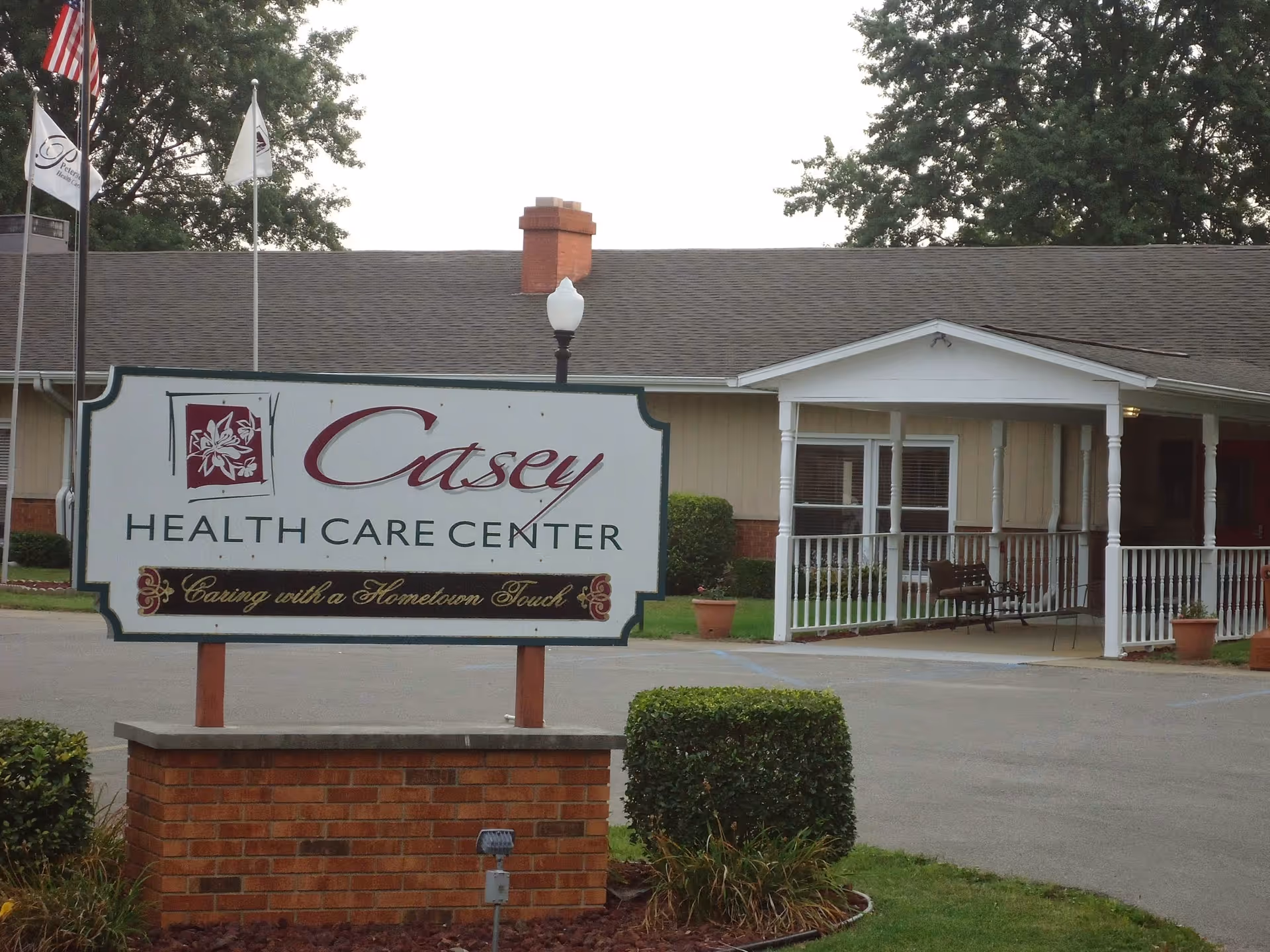 Exterior view of Casey Health Care Center showing a large sign with the facility's name and slogan 'Caring with a Hometown Touch' in front of a single-story building with a covered entrance and a bench.