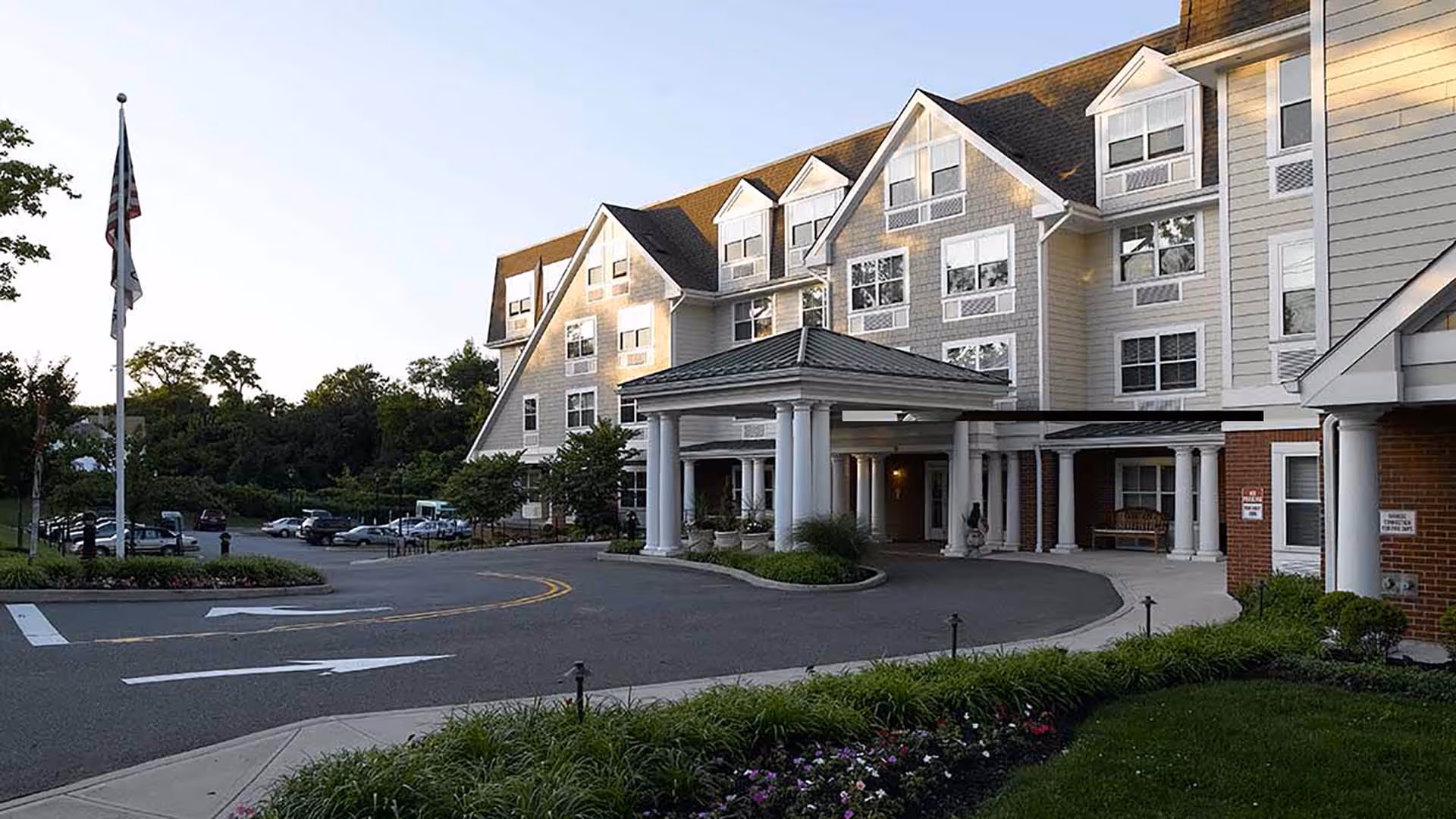 Exterior view of a senior living facility named Atria Tanglewood featuring a multi-story building with a covered entrance supported by white columns, surrounded by greenery and a parking area with several cars.
