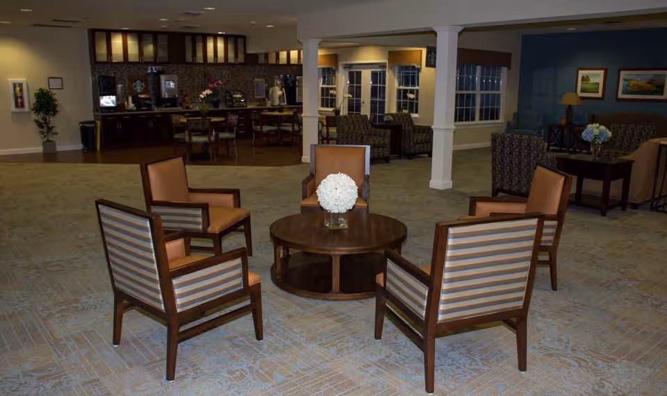 A spacious common area in a senior living facility featuring a round wooden table with a white floral centerpiece surrounded by four chairs with striped and solid upholstery. In the background, there is a kitchenette area with cabinets, a coffee machine, and additional seating. The room has carpeted flooring, large windows, and decorative columns.