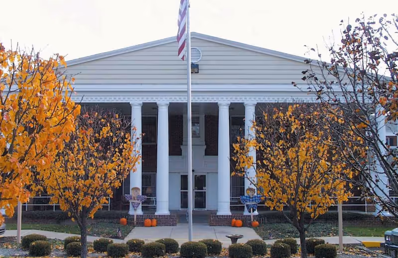 Front exterior view of a building with white columns and a triangular pediment. There are trees with orange autumn leaves on either side of the walkway leading to the entrance. An American flag is raised on a flagpole in front of the building. Small pumpkins and scarecrow decorations are placed near the entrance.