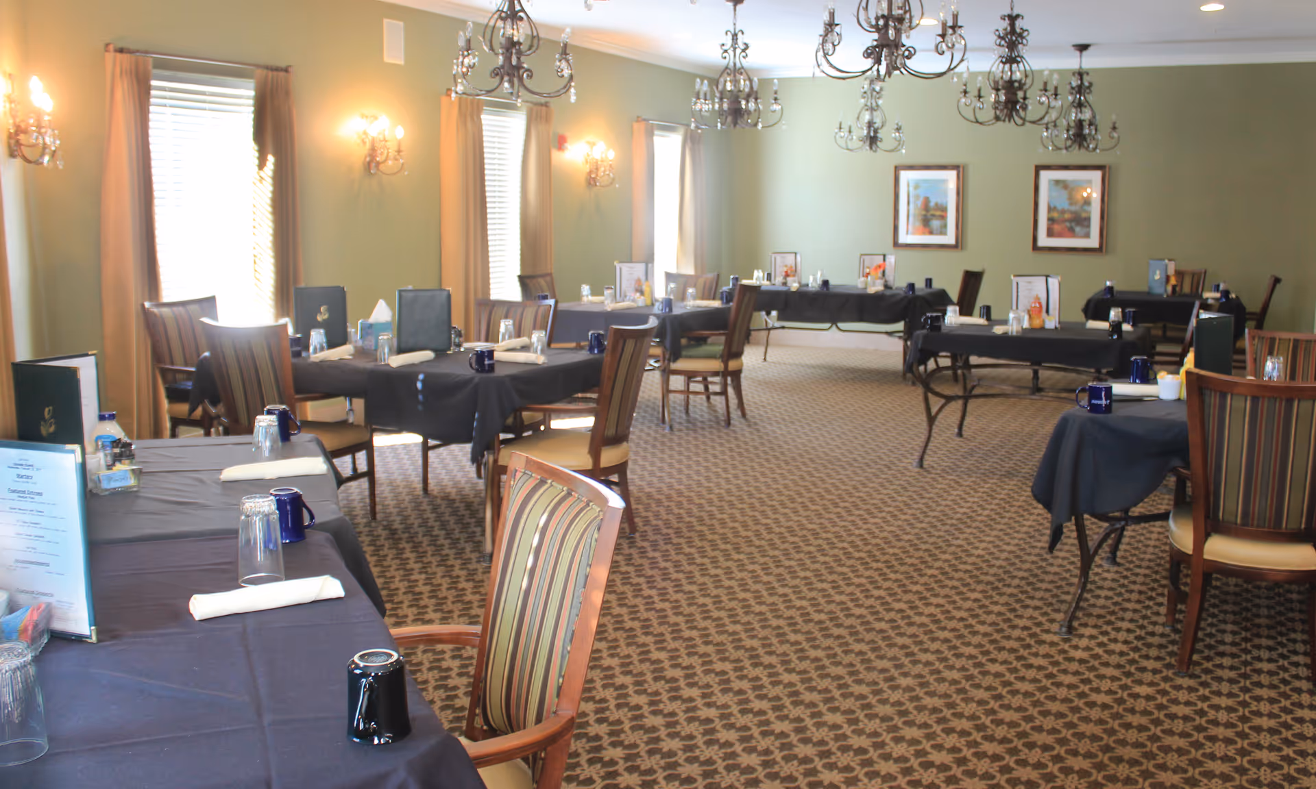A dining room with several tables covered in black tablecloths, each set with menus, napkins, cups, and glasses. The room has patterned carpet, green walls with framed pictures, multiple windows with curtains, and ornate chandeliers hanging from the ceiling.