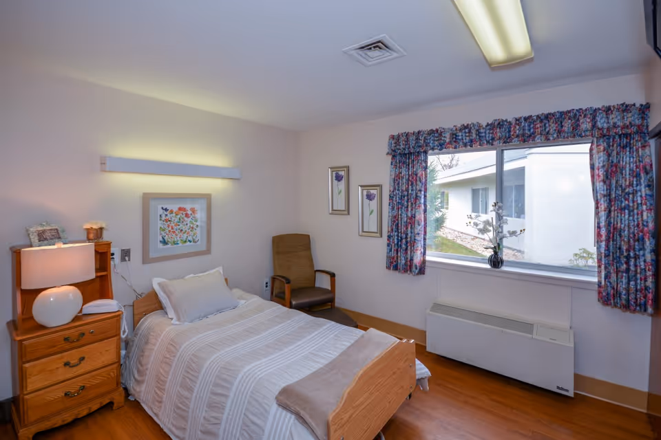 A small, neatly arranged bedroom with a single bed, bedside table and lamp, a chair, and a window with floral curtains.