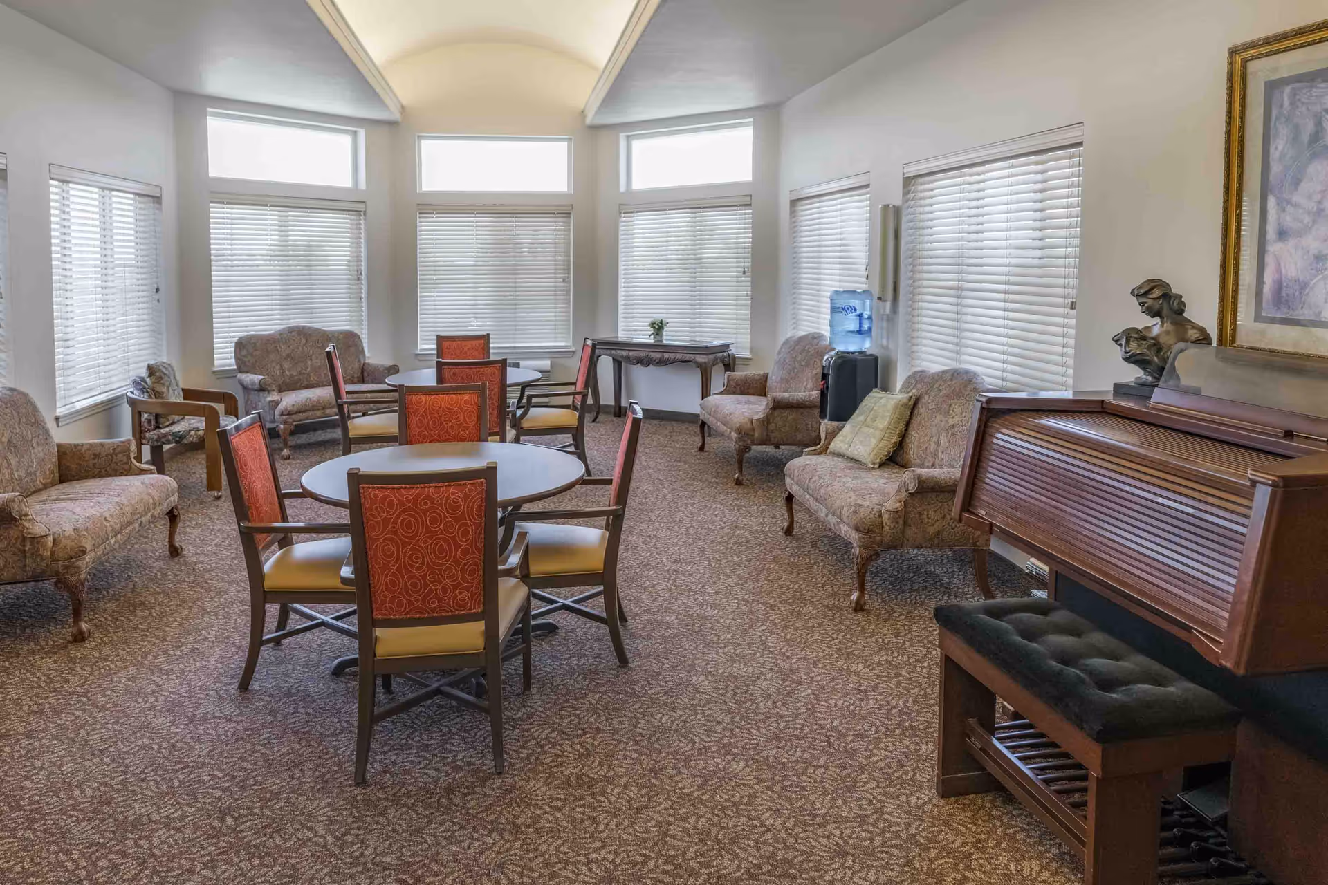Bright communal sitting room with round tables, upholstered chairs and armchairs, a piano, and large windows.