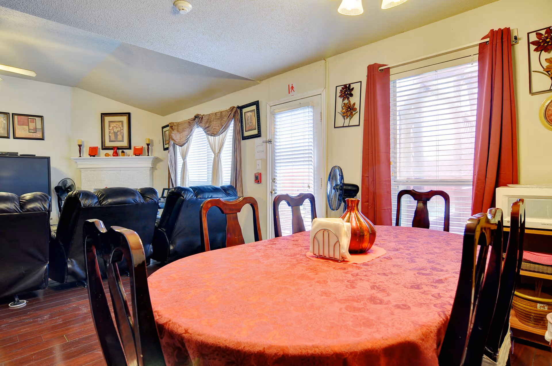 Interior view of a residential care home living and dining area featuring a round dining table covered with a red tablecloth, surrounded by wooden chairs. In the background, there are black leather recliners facing a white fireplace with framed artwork above it. The room has windows with blinds and red curtains, and a door with a window covered by blinds.