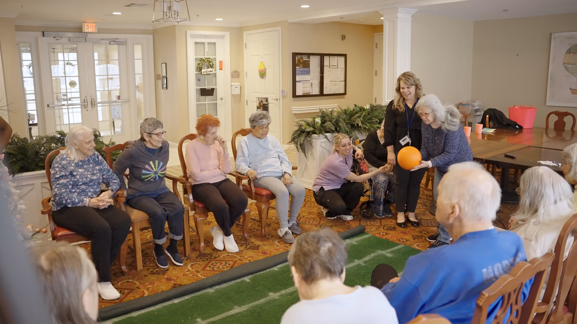 A group of elderly residents seated in a circle in a well-lit common room at a senior living facility, participating in a group activity involving an orange ball, assisted by two staff members.